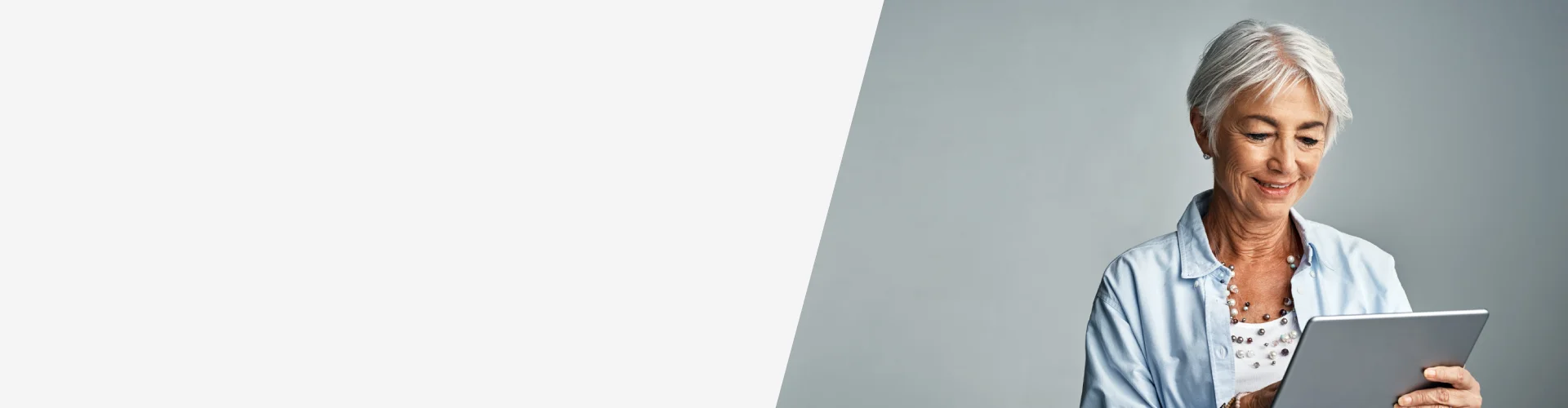 Persona mayor usando una tablet sobre fondo gris, vestida con camisa clara y collar