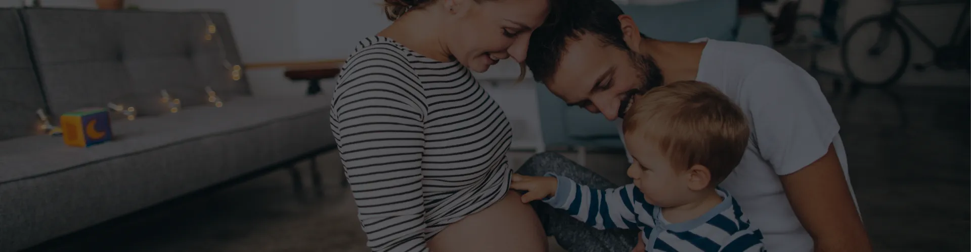 Familia en casa interactuando con el vientre de una mujer embarazada, sentados en el suelo junto a un sofá