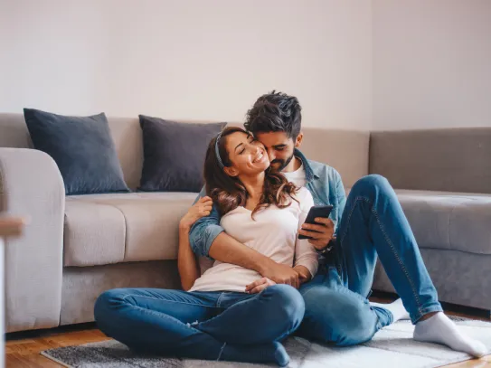 Pareja sentada en el suelo apoyada en un sofá, mirando un teléfono móvil en un salón hogareño