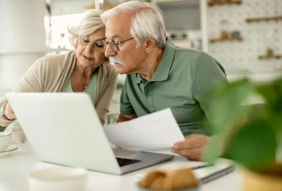 Dos personas mayores sentadas en una mesa blanca, consultando información en un ordenador portátil. Una de ellas sostiene un documento. 