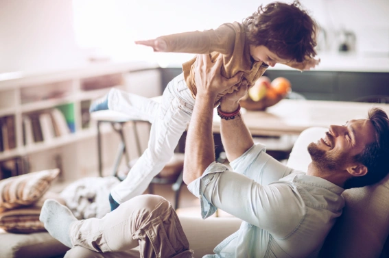 Persona adulta levantando a un niño en el aire mientras juegan en una sala de estar luminosa con sofá y estantería al fondo