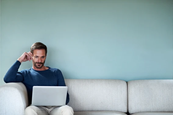 Persona sentada en un sofá usando un ordenador portátil sobre sus piernas, con fondo de pared lisa en tono verde azulado.