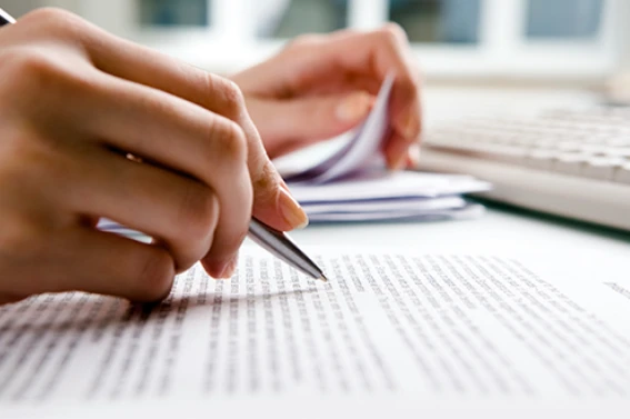 Mano escribiendo sobre documento impreso con bolígrafo, revisión de contrato legal junto a teclado en escritorio de oficina