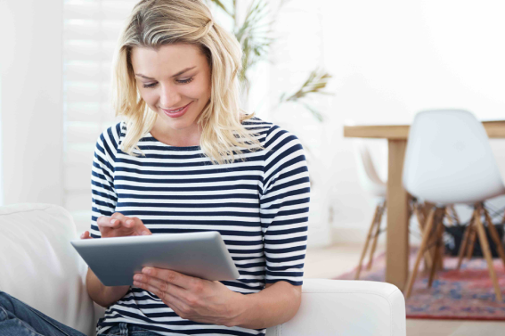 Persona sentada en un sofá usando una tablet en un entorno doméstico moderno, con luz natural y mobiliario minimalista al fondo.