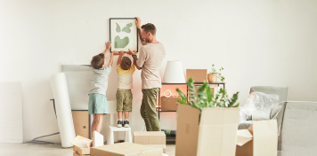 Persona colocando un cuadro en la pared junto a dos menores en una habitación con cajas, concepto de mudanza y seguros de hogar.