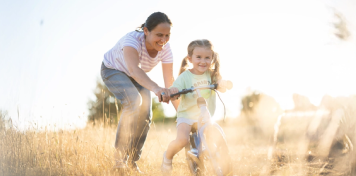 Adulto ayudando a una niña a montar en bicicleta en un campo soleado, concepto de protección y seguros de vida.