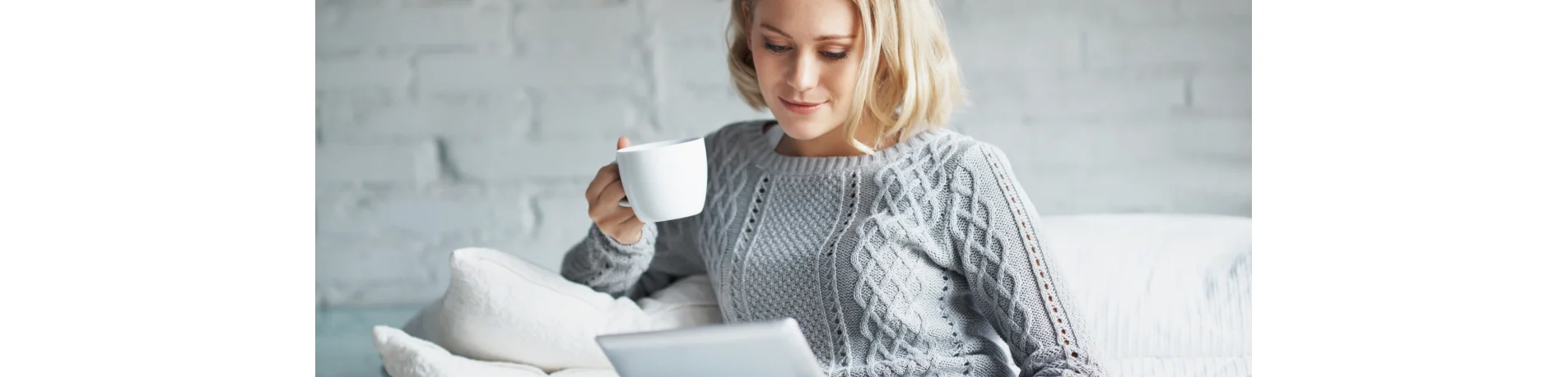 Persona sentada en un sofá con jersey gris, sosteniendo una taza de café y usando una tablet en un ambiente acogedor