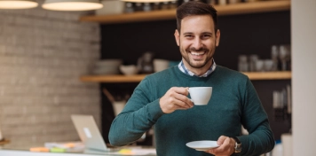 ersona en cocina moderna sosteniendo una taza y un plato, con portátil abierto sobre la encimera