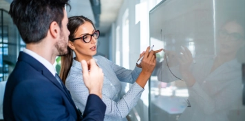 Dos personas en oficina escribiendo en una pizarra con gráficos, colaborando en análisis financiero
