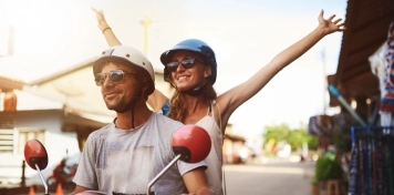 Dos personas montando en motocicleta sobre fondo urbano, con cascos y brazos extendidos hacia arriba en gesto dinámico