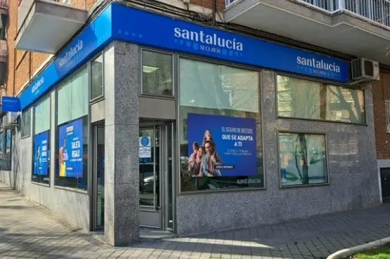 Fachada acristalada de una oficina de Santalucía Seguros en una esquina urbana, con rótulos corporativos azules y carteles promocionales en los ventanales